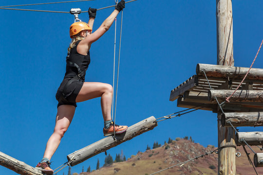 Adventure climbing rope park - a young woman walks along logs and ropes at a height against the background of mountains and blue sky. training mountaineers in the mountains. sports streets