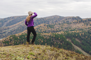 Fototapeta premium A girl in a lilac jacket looks out into the distance on a mountain, a view of the mountains and an autumnal forest by an overcast day. Free space for text