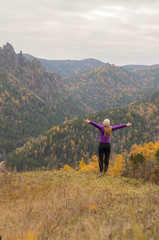 A girl in a lilac jacket jumps on a mountain, a view of the mountains and an autumnal forest overcast. Free space for text