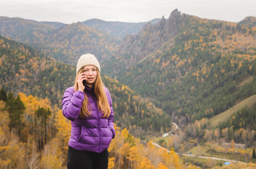 Fototapeta na wymiar A girl in a lilac jacket talking on the phone in the mountains, an autumn forest with a cloudy day, free space for text