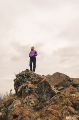 A girl in a lilac jacket looks out into the distance on a mountain, a view of the mountains and an autumnal forest by an overcast day, free space for text
