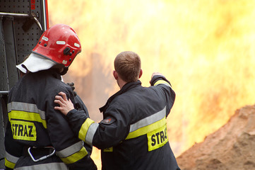 Two firefighters in dark uniforms with high-visibility yellow reflective stripes and the word "STRAŻ" (Polish for "Guard" or "Fire Service") on their backs, standing near a large fire. © Jacek