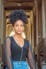 Fototapeta na wymiar Young beautiful African American Woman with afro hairstyle wearing mesh sheer long sleeve shirt blouse, walking on narrow vintage street in New York, listening music with earphone and cell phone..