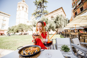 Young woman in red dress photographing sea Paella, traditional Valencian rice dish, sitting outdoors at the restaurant in Valencia