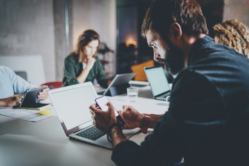 Bearded young coworker working at night office.Man using contemporary laptop and typing on modern smartphone.Horizontal.Blurred background.
