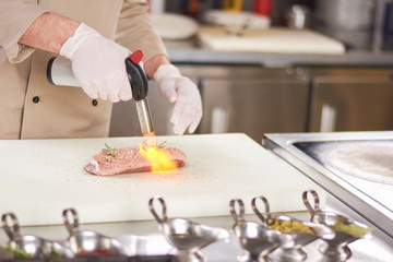 Fototapeta na wymiar Chef hands with torch burner. Torch blowing fire on meat. Chef is preparing duck breast.
