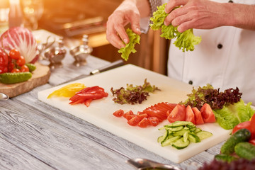 Chef preparing food at professional kitchen. Male chef holding in hands green lettuce salad. Cutting vegetables on white board.