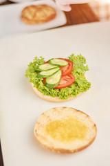 Fototapeta na wymiar Cooking of hamburger at kitchen. Burger with lettuce, tomatoes and fresh cucumbers on table. Burger preparing at cafe.