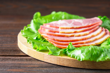 Sliced ham with fresh green lettuce leaves on a round cutting board. Meat products on a brown wooden table.