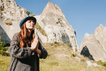 Naklejka na ściany i meble Bottom view - beautiful young woman in hat and gray sweater meditates with folded hands and closing eyes against backdrop of beautiful mountains and hills on sunny day