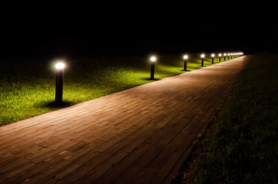 Night landscape of the park. The path is covered with beige tiles and is lit by flashlights. Around the path there is a lawn with green grass.