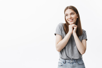 Close up of charming good-looking young student girl with dark hair and eyes in casual outfit smiling, looking aside with dreamy and happy face expression, holding hands together under face.