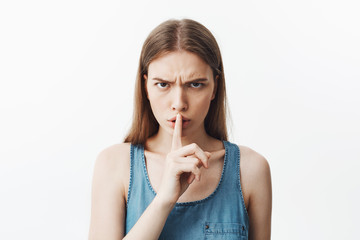 Close up of good-looking young caucasianstudent girl with dark hair in blue shirt holding hand in fromt of lips, making hush gesture with serious and unsitisfied expression. Woman tries to calm down