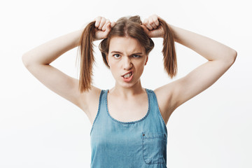 Fototapeta premium Isolated portrait of funny youn caucasian woman with dark hair in bright blue shirt holding hair with hands, looking in camera with silly face expression, showing teeth, posing for funny photo on