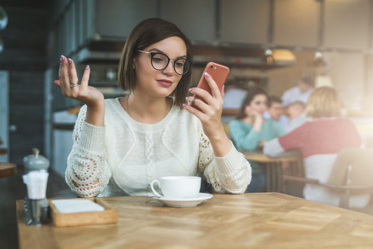 Young businesswoman is sitting in coffee shop at table and using smartphone, working. E-learning,online marketing,education. Hipster girl looking at phone screen in surprise, checking email, chatting.