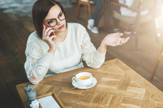 Young businesswoman in glasses and white sweater is sitting in cafe at wooden table and talking on cell phone. Telephone conversations. Hipster girl freelancer discusses working questions over phone.