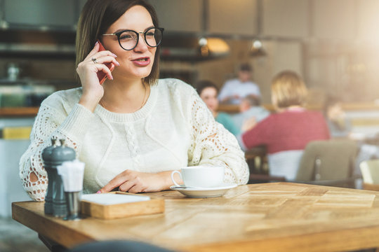 Young businesswoman in glasses and white sweater is sitting in cafe at wooden table and talking on cell phone.Telephone conversations.Hipster girl is talking on phone with her friend.Instagram filter.