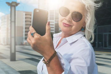 Summer day. Middle-aged woman, a pensioner, dressed in a white shirt and sunglasses, stands on a city street and uses a smartphone. In the background is a modern glass building. Social media, network.