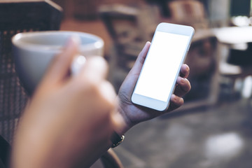Mockup image of woman's hands holding white mobile phone with blank screen while drinking coffee in modern loft cafe
