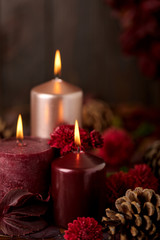 Three candles of crimson and pink color on a dark background with cones, leaves and daisies.