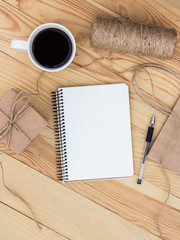 A blank notebook laying on wooden table. It is surrounded by  black pen, cup of coffee, present, rope twine and a paper bag.