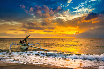 A fishing boat goes to the ocean in the evening. Bright colorful orange sun hidden in dense blue clouds over the Bali Sea. A white wave pours a foam to the sandy beach. Sunset on the Lombok Indonesia