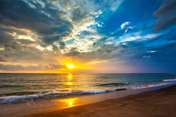 A bright colorful orange sun makes the rays through dense blue clouds on the Bali Sea. View from the sand beach. Sunset on the Lombok island, Indonesia.
