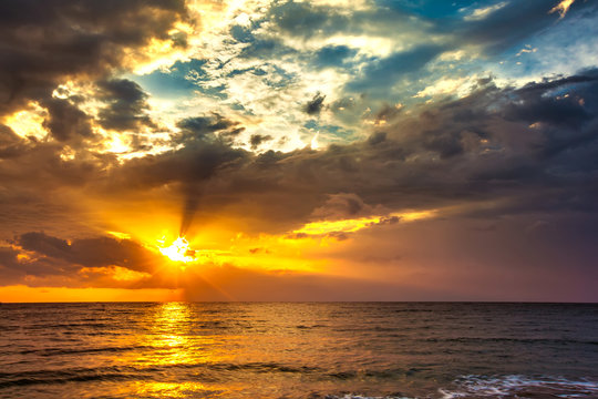 A bright colorful orange sun makes the rays through dense blue clouds on the Bali Sea. Sunset on the Lombok island, Indonesia.