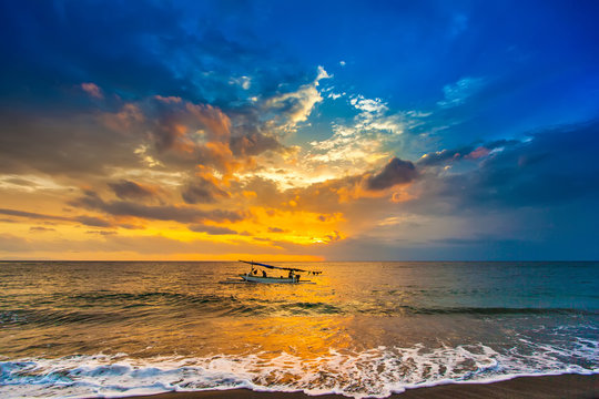 A fishing boat goes to the ocean in the evening. Bright colorful orange sun hidden in dense blue clouds over the Bali Sea. A white wave pours a foam to the sandy beach. Sunset on the Lombok Indonesia