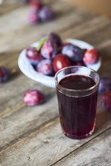 Glass of plum juce with fresh fruits in plate on wooden background