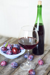 Glass of plum wine with bottle and fresh fruits in plate on wooden background