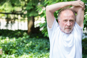 Closeup portrait, senior mature man in white shirt, stretching arms, isolated green trees background. Warming up