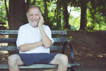 Fototapeta premium Closeup portrait, older, happy ecstatic man sitting on bench talking on cell phone, isolated outdoors background