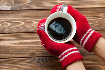 Hands in red knitted gloves holding a hot cup of coffee on the wooden background. Concept of autumn time, warmth and coziness. Instagram filter