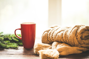 Christmas Hot Coffee in red cup on wooden table on a frosty winter day window background with knitted scarf and christmas tree branches. Home cozy holidays background.