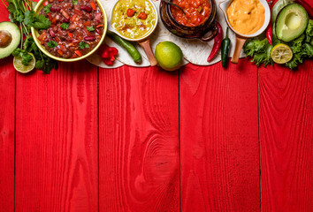 Vegetarian Mexican food concept: refried black and red beans. guacamole, salsa, chili, tortilla chips and fresh ingredients over vintage red rustic wooden background. Top view