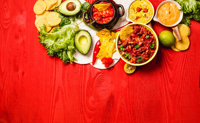 Vegetarian Mexican food concept: refried black and red beans. guacamole, salsa, chili, tortilla chips and fresh ingredients over vintage red rustic wooden background. Top view