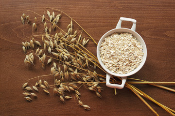 Oat ears stems and oat flakes in a white pot on a dark brown wood background. Top view. oat flakes small size grind. Useful fiber-rich product. Dietary breakfast from healthy foods