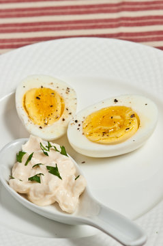 Halved hard boiled egg with fresh ground pepper on a round, white plate and a ceramic spoon of mayo and chopped parsley on a red and white stripped cloth-Paleo diet