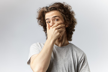 Frightened and astonished young male with shaggy hair covering mouth while accidentally eavesdropping some gossip or shocking unexpected news, restraining himself, trying hard to keep silent