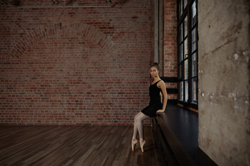 Portrait of elegant slim young Russian ballerina in black pack, white pantyhose and ballet shoes sitting on wide windowsill in studio and relaxing during intense exercise, preparing for performance © Anatoliy Karlyuk