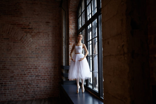 Graceful professional young woman ballet dancer standing on pointes on windwosill at lrage window dressed in white tutu while training in studio with blank copy space wall for your promotional content