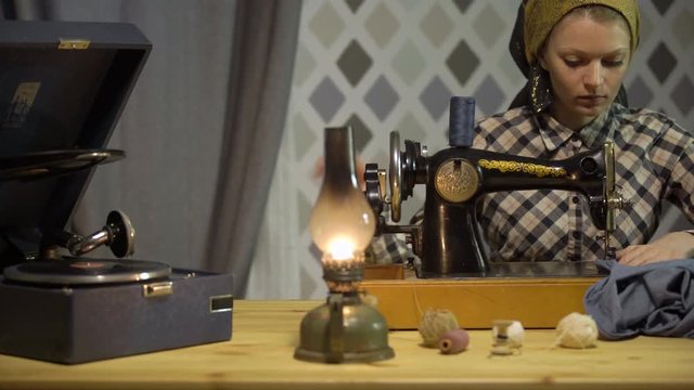 Retro seamstress girl sews cloth with old manual hand sewing machine. Woman works at home or workshop at night with kerosene lamp, listens music vinyl plate, gramophone or phonograph