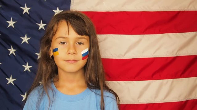 A little girl in a boxing rack with the Ukrainian and Russian flag. A child with a Russian and Ukrainian flag on his face against the background of the flag of America.