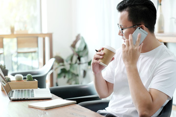 Young asian man talking smart phone and holding a coffee cup while working with laptop computer in cafe, working in casual lifestyle