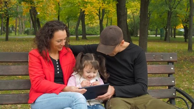 Happy family with gadget in the park. Parents with a little daughter with a tablet in an autumn park.