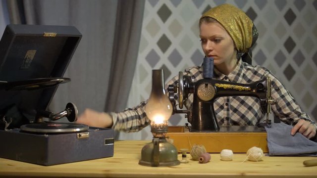 Tailor girl sews cloth with old manual hand sewing machine. Retro seamstress woman works at home or workshop at night with kerosene lamp, listens music vinyl plate, gramophone or phonograph