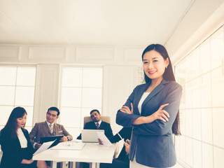 Confident young Asian businesswoman standing with folded arms smiling at the camera in a boardroom with male and female colleagues background.  Brainstorm meeting.  Business successful conceptual.