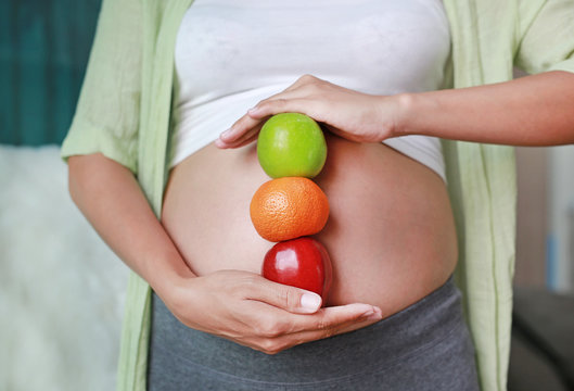 Pregnant woman holding Green-Red Apple and Orange fruit at her belly.