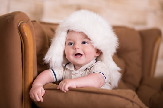 Happy newborn child baby boy with pink cheeks posing on hege retro casual style brown couch divan sofa with big white fur winter warm hat